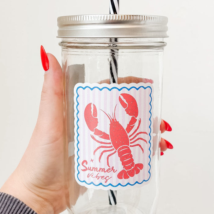 Hand holding mason jar tumbler up close that reads summer vibes in red ink with lobster design on pale pink and white striped background with blue border
