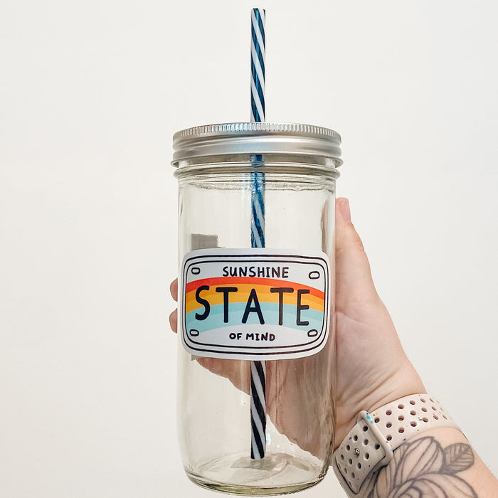 Hand holding mason jar that reads sunshine state of mind in the shape of a license plate with a rainbow across the middle.