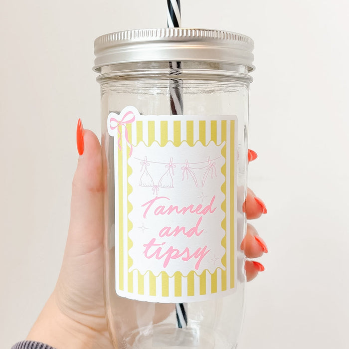Hand holding mason jar tumbler up close that reads tanned and tipsy in pink font with yellow striped background and pink bow design