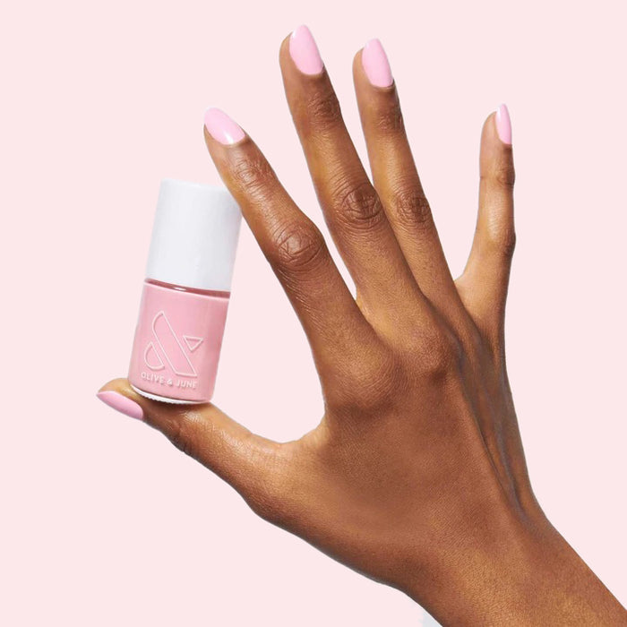 Hand with light pink manicure holding Olive and June nail polish from The Pink Mama Set against soft pink background