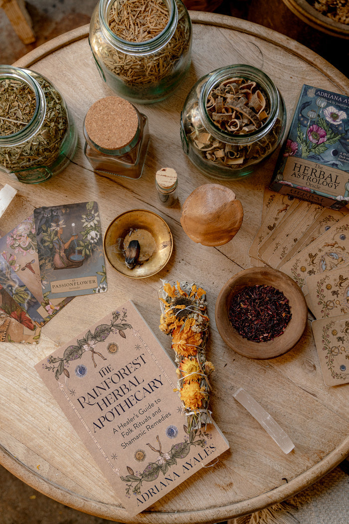 A styled flat-lay of a 'The Painforest Herbal Apothecary' book by Adriana Walls, surrounded by glass jars of dried herbs, a sage bundle, a crystal ball, and cards on a rustic wooden table.