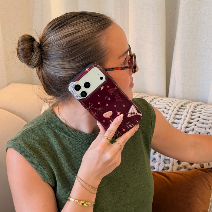 Woman in a dark green jumper with a white lace shirt underneath holds a burgundy chocolate brown phone case with decorative beaded illustrations on it, such as a bow, a strawberry, a cocktail, stars and cherries.