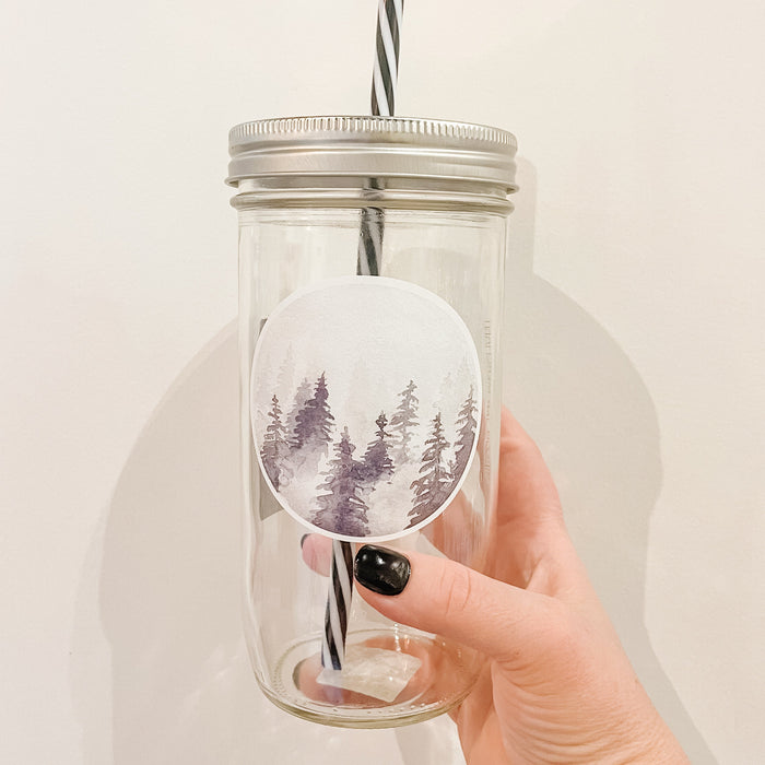 Hand holding a mason jar tumbler that has a circle decal of trees in different shades of grey in fog.