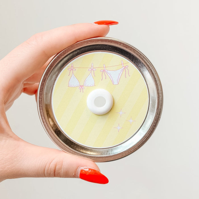 Hand holding mason jar patterned straw lid up close that has a pink bikini on a clothesline design on light yellow striped background