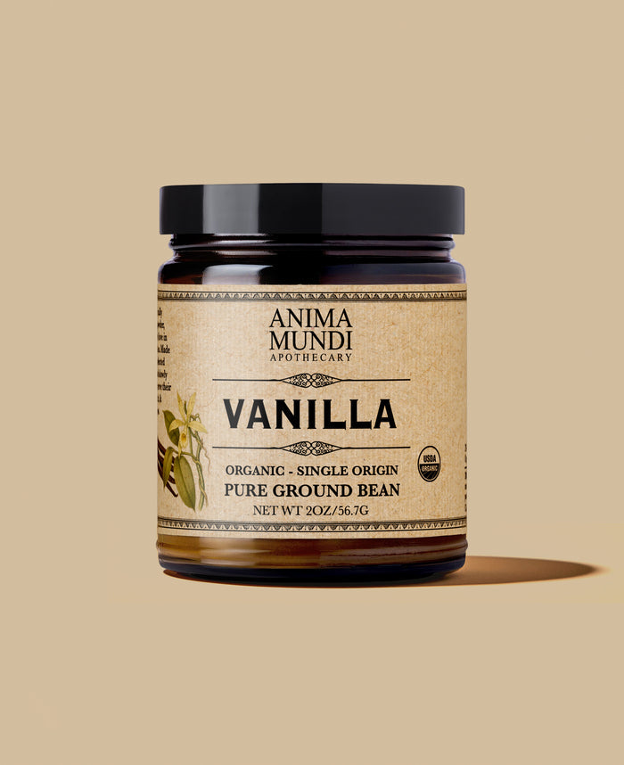 A jar of Anima Mundi Apothecary Vanilla Pure Ground Bean on a solid beige background. The product is in a dark amber glass jar with a black lid and a beige label featuring black text and a vanilla bean illustration.