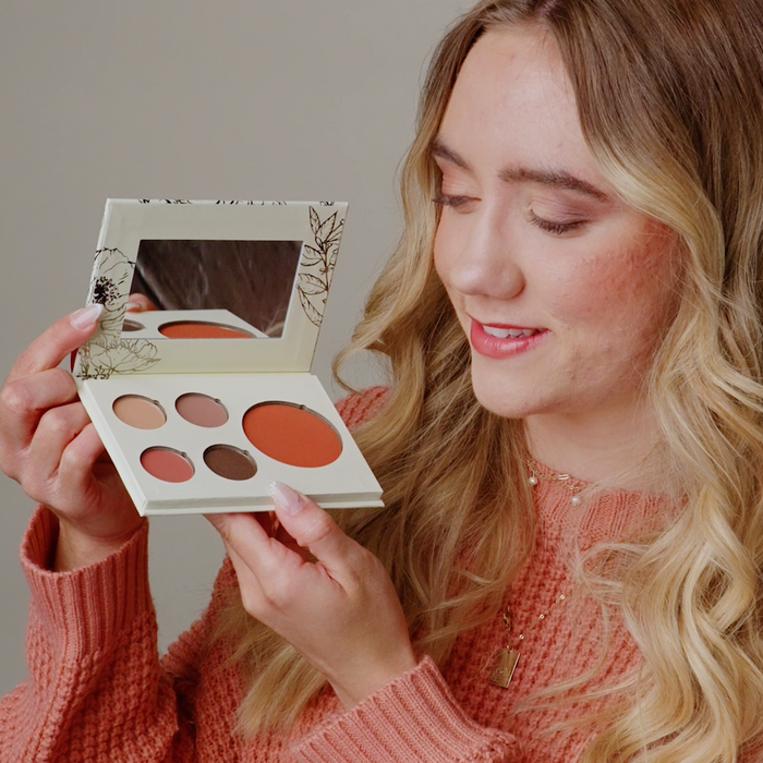 A woman with blonde hair wearing a textured peach sweater holds an open makeup palette with five round pans in warm neutral and peach shades against a plain beige background.