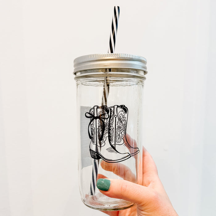 Hand holding a mason jar tumbler with a design that has western boots with a bow on the side in black ink and clear background