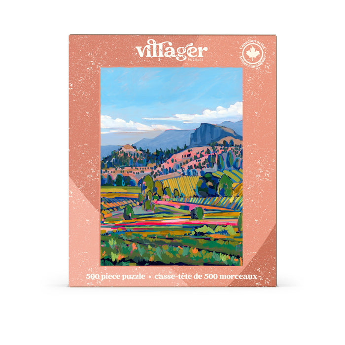 The Villager Puzzles “Wine Country | 500 Piece Jigsaw Puzzle” features a vibrant painted landscape of fields, hills, trees, and distant mountains beneath a blue sky, with artwork inspired by Amy Dixon.