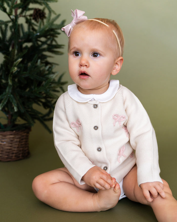 Viverano Organics baby cardigan in cream with pink bow embroidery worn by seated baby girl