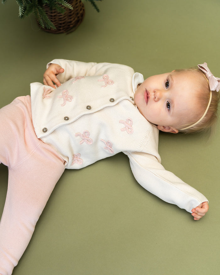 Viverano Organics baby cardigan in white with pink bow embroidery worn by baby lying on green surface